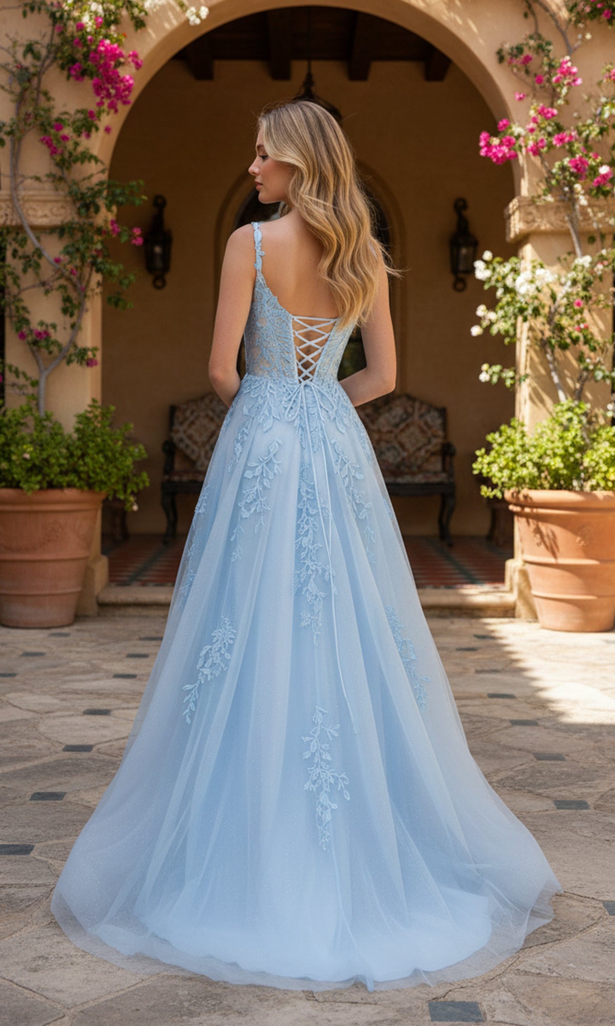 Wearing the Long Prom Dress: PromGirl Exclusive 250261, a woman stands outdoors with her back to the camera, framed by a stone archway and potted flowers. The floor-length gown features light blue lace applique.